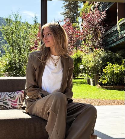 Donna seduta su divano da esterno in giardino curato, con completo beige e t-shirt bianca, illuminata da luce naturale. Atmosfera elegante e rilassante tra verde e fiori colorati.