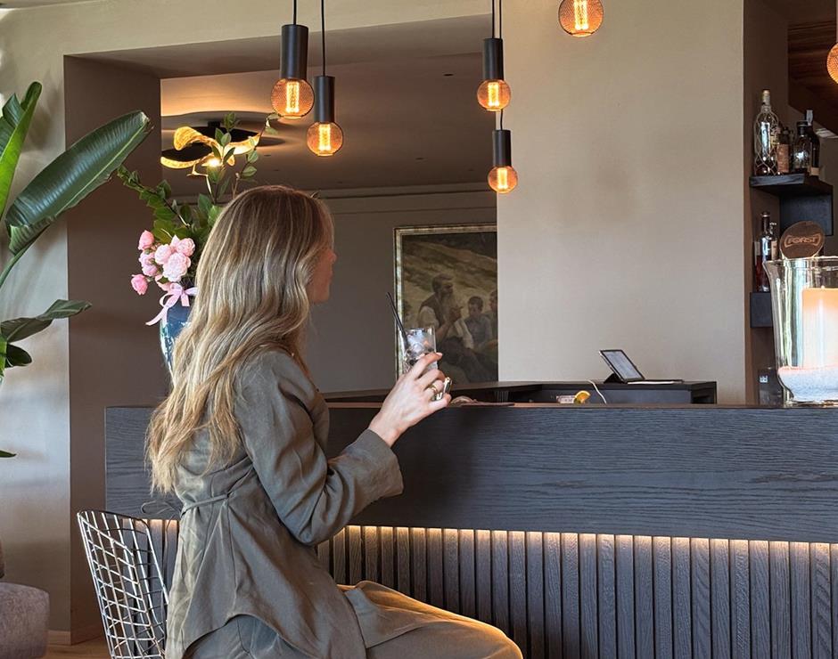 Donna elegante seduta al bancone bar con drink in mano, illuminazione calda e design moderno. Atmosfera raffinata e accogliente in ambiente contemporaneo.