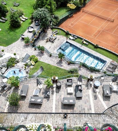 panoramica dall’alto dello Sport Hotel Panorama di Fai della Paganella con piscina esterna riscaldata, vasca idromassaggio e ampio solarium immerso nel verde. Accanto, campo da tennis in terra rossa