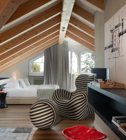 Elegante suite mansardata allo Sport Hotel Panorama di Fai della Paganella, caratterizzata da soffitto con travi in legno a vista e ampie finestre che donano luce naturale agli ambienti. Arredi contemporanei, dettagli di design e atmosfera accogliente