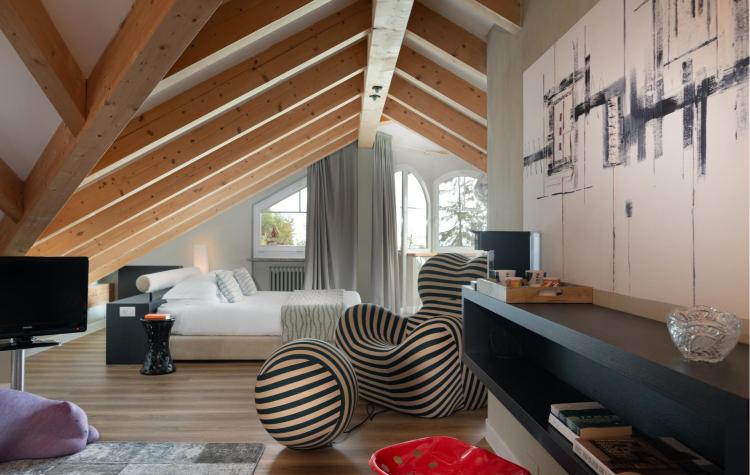Elegante suite mansardata allo Sport Hotel Panorama di Fai della Paganella, caratterizzata da soffitto con travi in legno a vista e ampie finestre che donano luce naturale agli ambienti. Arredi contemporanei, dettagli di design e atmosfera accogliente